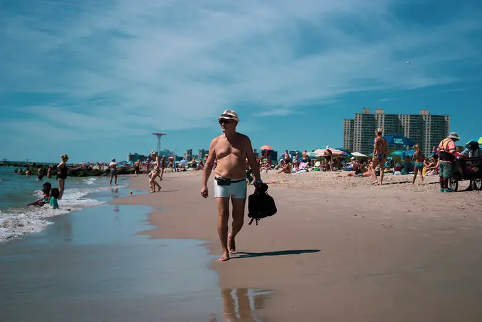 A man wearing white swim trunks and a white hat walks along a crowded beach in Coney Island, Brooklyn.